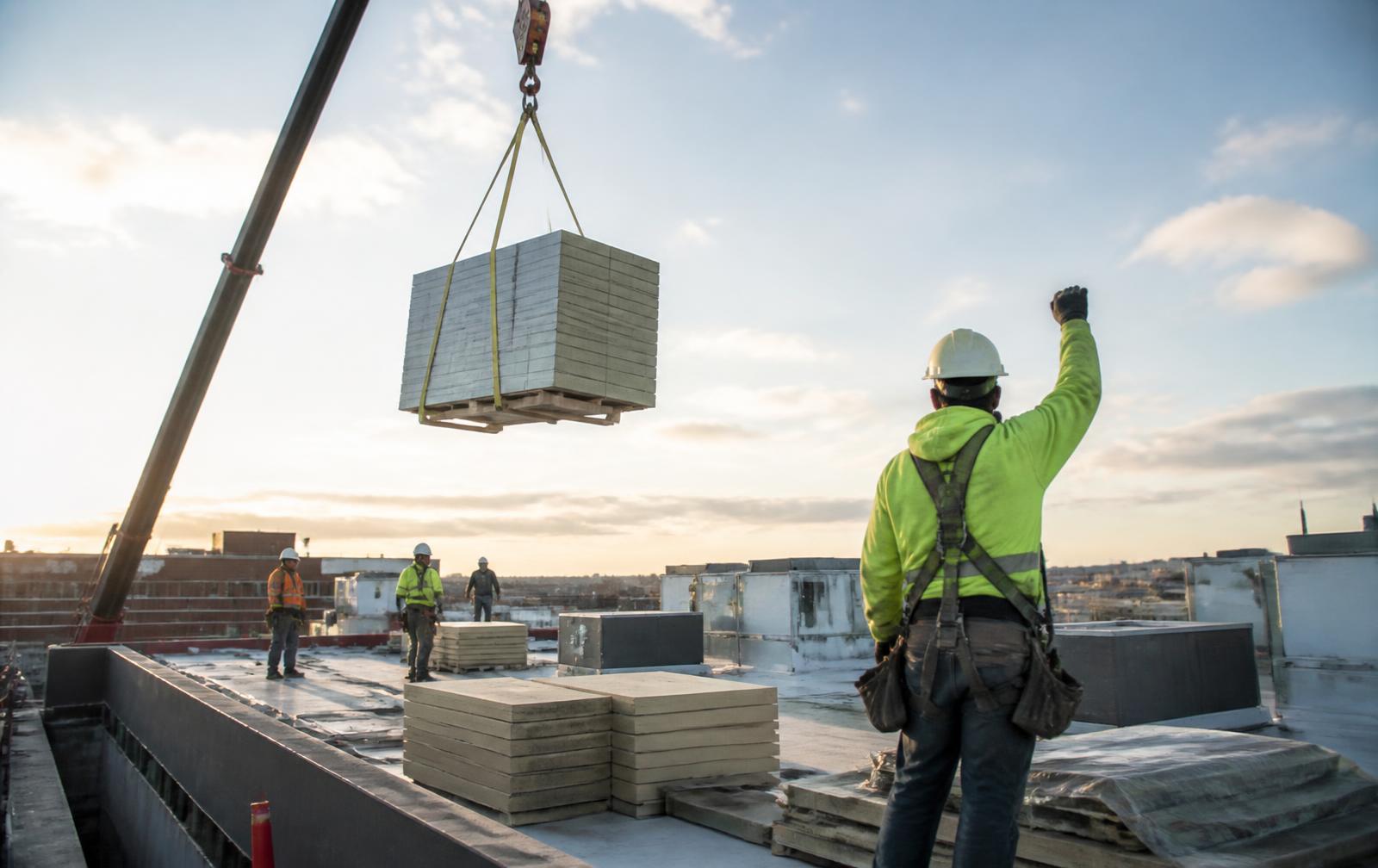 Roofing crew staging insulation board on a commercial rooftop while a crane hoists materials